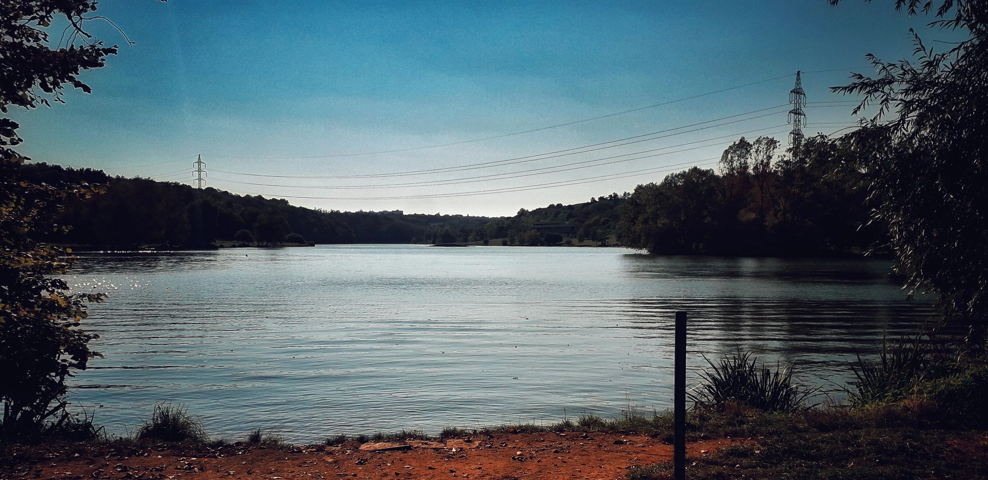 Široký pohled na řeku/jezero, modrá obloha, v dálce stožáry a břehy se zelení
