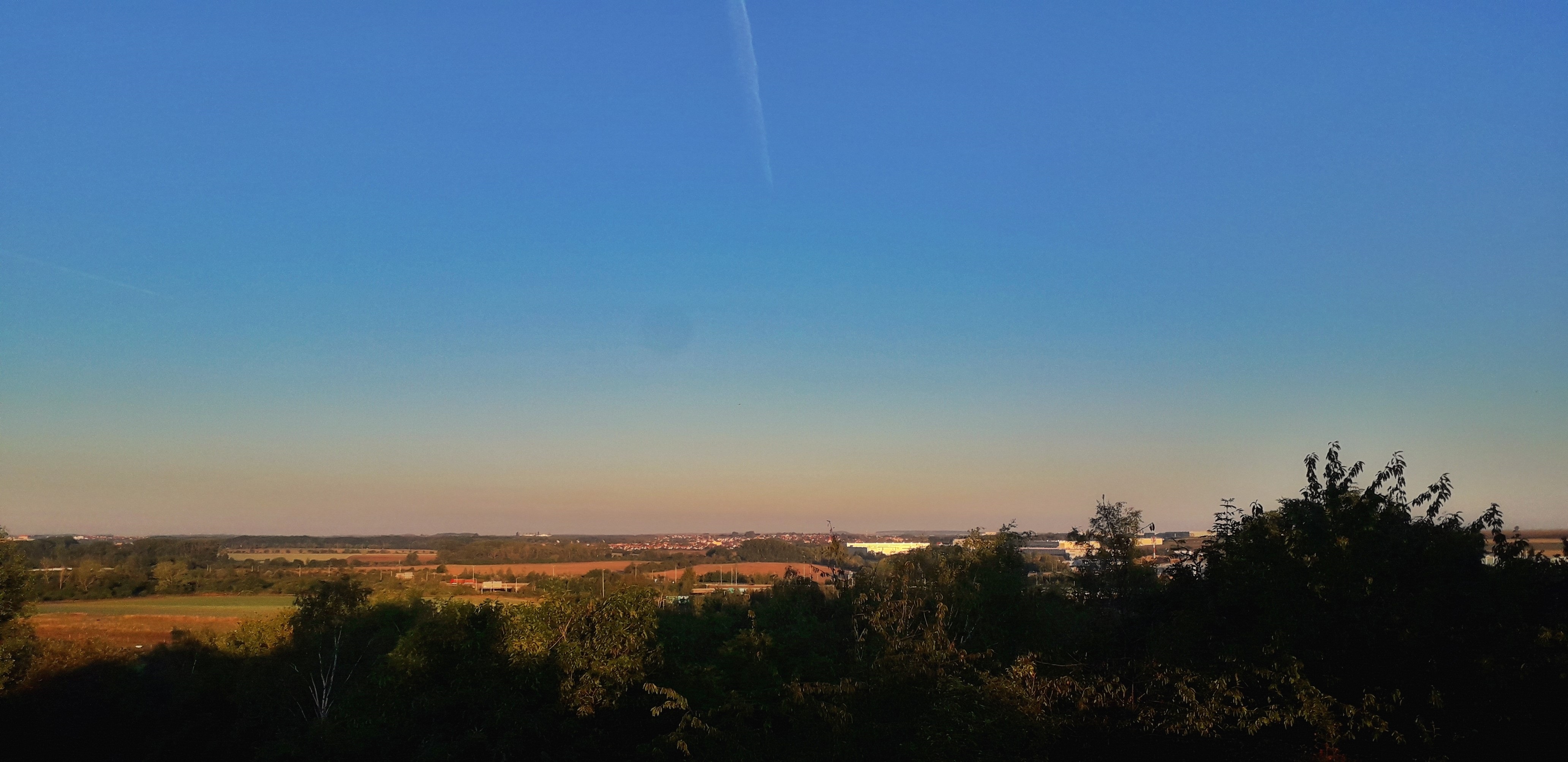 Pohled přes pole a zeleň k vzdálenému městu pod čistou modrou oblohou