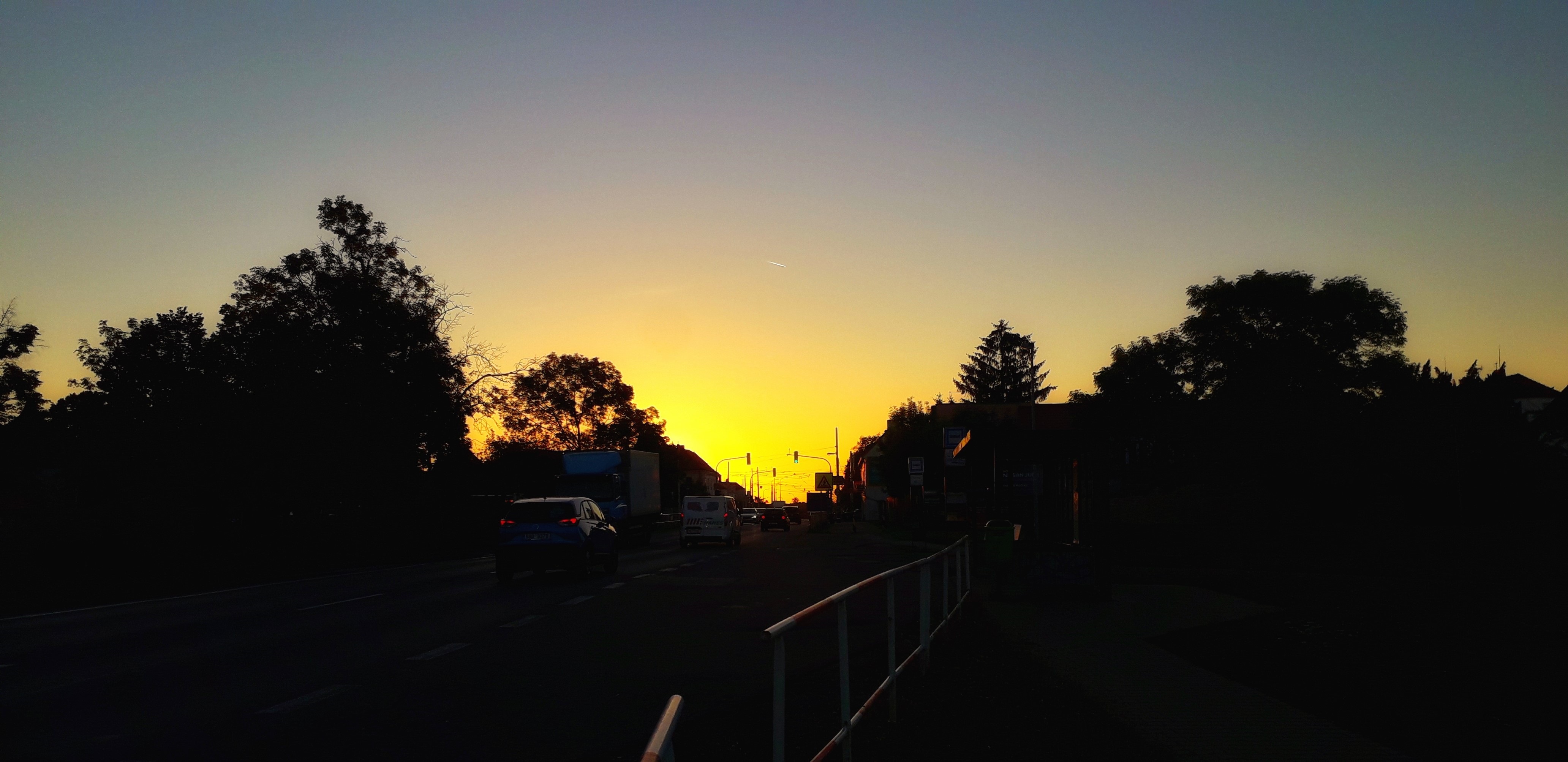 Západ slunce nad ulicí; tmavé siluety stromů a aut proti oranžové obloze